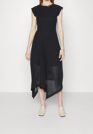 Robe noire sans manches avec un corsage ajusté et un ourlet asymétrique, en tissu léger. Le modèle est assorti à des chaussures beige à enfiler.