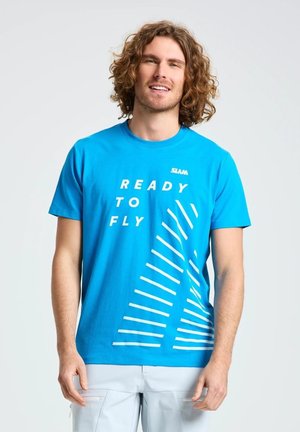 Un hombre sonriente luce una camiseta azul brillante con el texto «Ready to Fly» y gráficos blancos, y unos pantalones cortos claros, sobre un fondo liso.