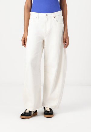 Hvite, vide denim-bukser med lommer og sydd sømmer, paret med svarte og brune sneakers med snøring og en teksturert såle.