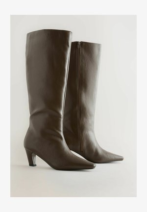 Bruine leren knie-hoge laarzen met een spitse neus, slanke vorm en een zijrits. De hakken zijn transparant en taps toelopend.