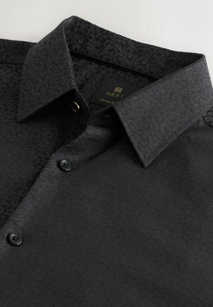 Must jätkuvalt must, tekstuuriga mustriga, struktureeritud krae, kaks nuppu ja sildiga. Materjal on sile ja pehme viimistlusega.