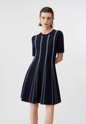 Donna che indossa un vestito nero a maniche corte con righe bianche verticali cucite, in piedi davanti a uno sfondo chiaro uniforme.