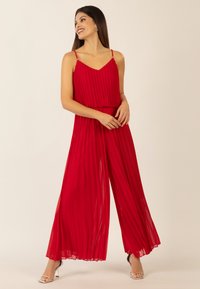 Roter, plissierter Jumpsuit mit dünnen, verstellbaren Trägern, einem tiefen V-Ausschnitt und einer fließenden, weiten Silhouette. Mit goldenen Akzenten an den Trägern.