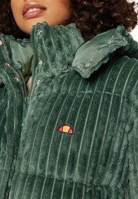 Groene corduroy pufferjack met ribbelstructuur, hoge kraag en geborduurd logo in rood en geel op de borst. Zilveren drukknoopsluitingen.