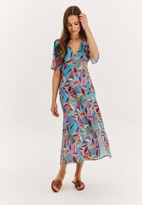 Maxi šaty z lehké, průsvitné tkaniny s barevným listovým vzorem. Mají výstřih do V a krátké rukávy, doplněné plochými sandály.