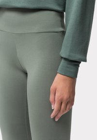 Legginsy w kolorze oliwkowym, o wysokim stanie, wykonane z miękkiego, elastycznego materiału o gładkiej strukturze. Posiadają dopasowany krój i subtelne detale szwów.