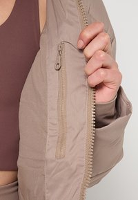 Ruka drži otvorenu bež podstavljenu jaknu kako bi otkrila unutrašnji zip zatvarač, osoba nosi smeđi top bez rukava ispod.