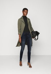 Casaco trench verde-oliva sobre um suéter preto de gola alta, combinado com jeans skinny azul-escuro e sandálias pretas. A segurar uma bolsa preta.