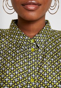 Gros plan d'une personne portant de grandes boucles d'oreilles créoles dorées et une chemise à col avec un motif géométrique vert et noir et des boutons jaunes.