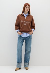 Giovane donna con capelli rossi che indossa un felpa marrone con mezza zip, una camicia azzurra a bottoni, jeans a gamba larga con risvolto e stivali beige in piedi.
