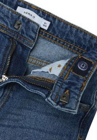 Jean en denim de couleur bleu foncé, avec des coutures visibles en orange, fermeture éclair et étiquette en tissu. Les éléments matériels notables incluent un bouton et un fermoir.