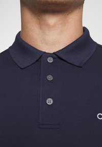 Navy polo shirt with a textured fabric, three gray buttons, and a classic collar. Subtle logo detail embroidered on the left side.