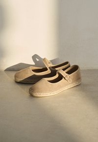 Beige espadrillosko med rundet tå, ruskindsstruktur, ankelrem og sål indpakket i jute, der hviler på en lys overflade.