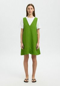 Vestido verde de lino en corte A con escote en V, llevado sobre una camiseta blanca. El diseño presenta un ajuste relajado y un estilo minimalista, sin patrones.