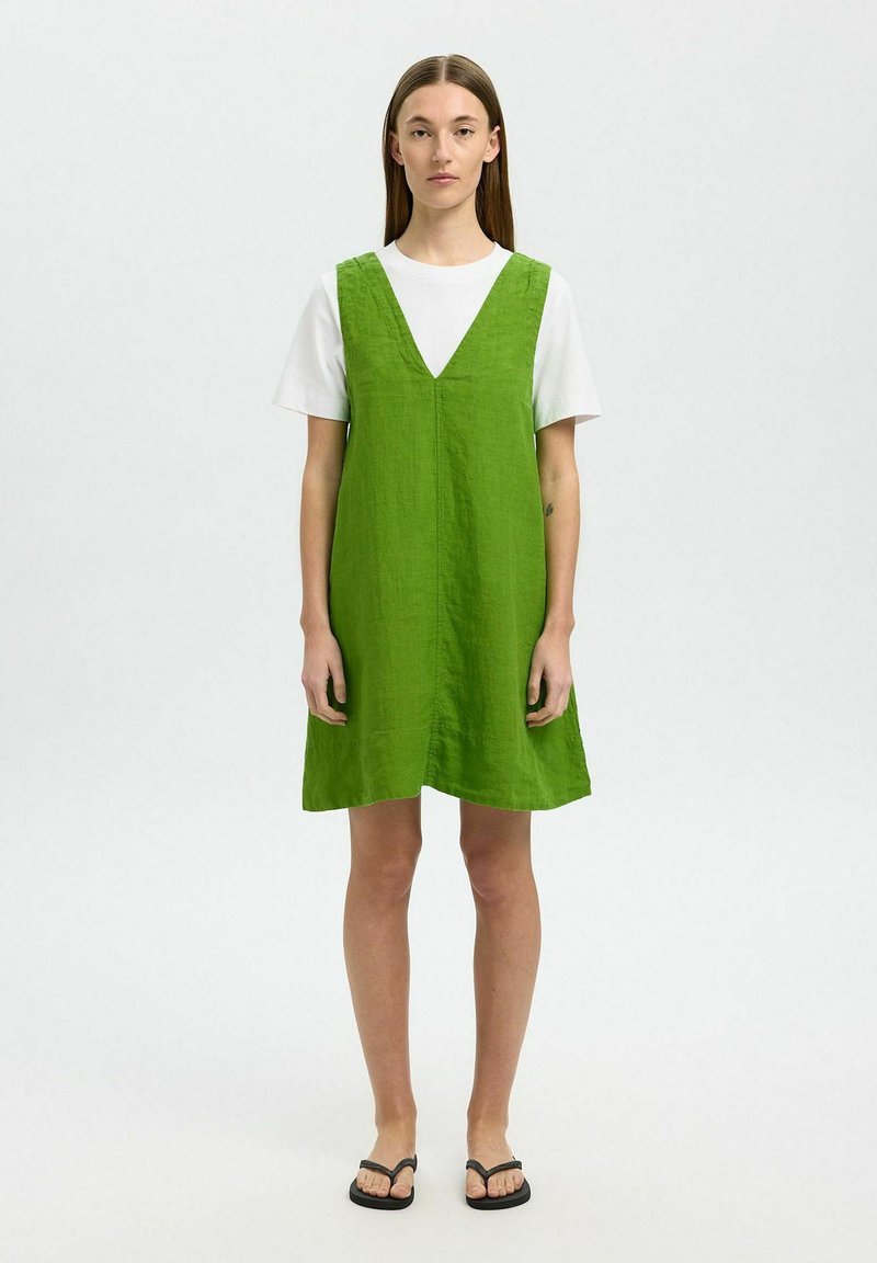 Vestido verde de lino en corte A con escote en V, llevado sobre una camiseta blanca. El diseño presenta un ajuste relajado y un estilo minimalista, sin patrones.