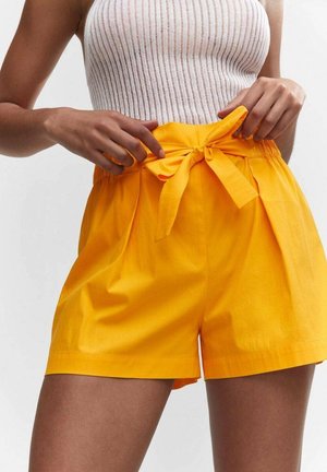 Femme portant un short taille haute jaune vif avec une ceinture nouée et un haut sans manches blanc côtelé, ajustant le short.