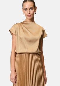 Blusa di seta beige con maniche corte, caratterizzata da morbide drappeggiature. Sotto, una gonna plissettata dello stesso colore crea una silhouette fluida e armoniosa.
