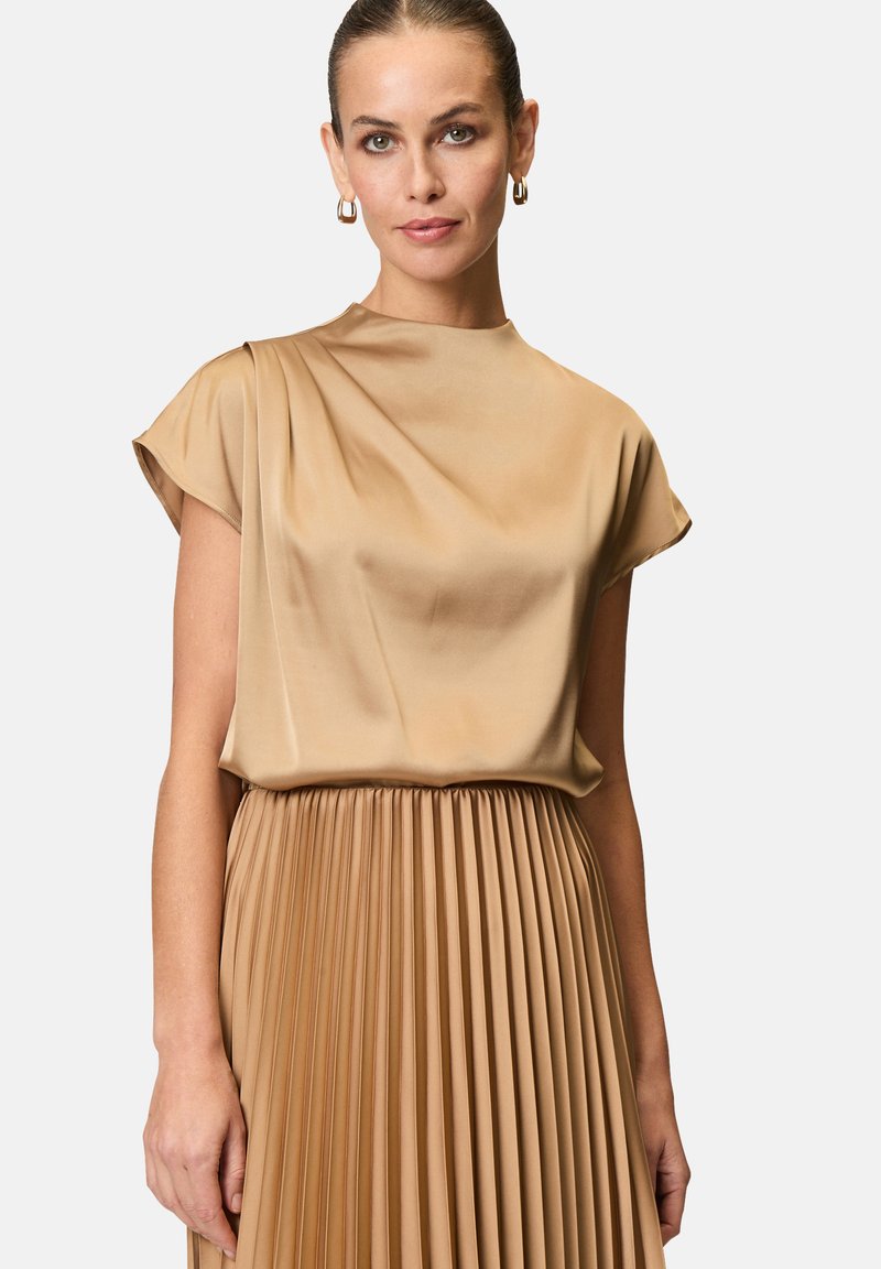 Blusa di seta beige con maniche corte, caratterizzata da morbide drappeggiature. Sotto, una gonna plissettata dello stesso colore crea una silhouette fluida e armoniosa.