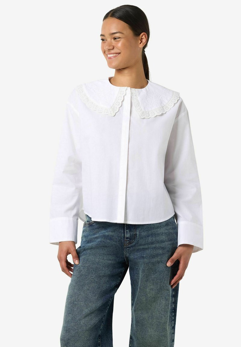 Biała krótka koszula z dużym, haftowanym koronkowym kołnierzem i długimi rękawami, wykonana z gładkiej bawełnianej tkaniny. Stylizowana z niebieskimi dżinsami z denimu.
