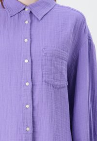Lavender button-up shirt made of textured fabric. Features a pointed collar, front pocket, and white buttons along the placket.