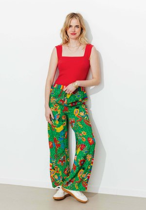 Femme blonde portant un haut rouge sans manches et un pantalon vert coloré à imprimés tropicaux, debout contre un mur blanc, chaussée de baskets blanches.