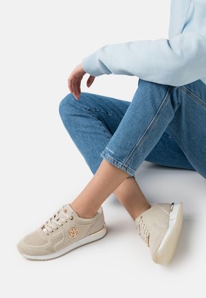 Jambes croisées portant un jean bleu et des baskets beige avec des accents de logo doré, sur un fond blanc.