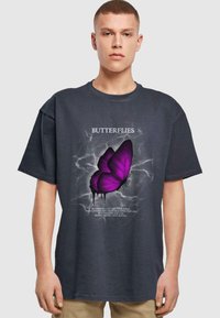 Donkergrijze oversized t-shirt met een centraal paarse vlinderafbeelding, witte tekst en een grijs abstract achtergrond.