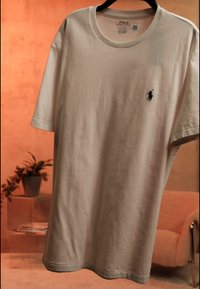 Beige Polo Ralph Lauren lyhythihainen t-paita, jossa musta hevosen logo rinnassa, ripustettuna mustalle naulakolle lämpimän sävyisen huoneen taustalla.