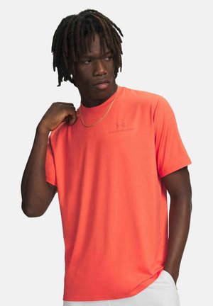 Uomo con dreadlocks che indossa una maglietta arancione vivace Under Armour e una catena d'oro, che guarda di lato con una mano vicino al collo.