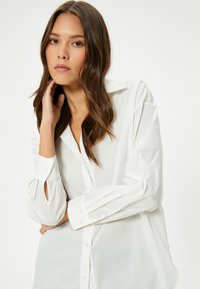 Camicia bianca con bottoni, maniche lunghe, caratterizzata da una vestibilità ampia, tessuto morbido e dettagli a fessura ai polsini delle maniche. Texture liscia.