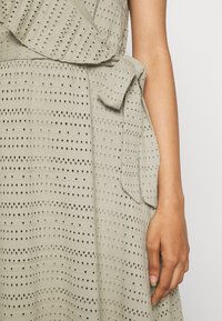 Světlé zelené šaty vyrobené z texturované látky s malými výřezy. Mají střih do zavinovacího stylu a pás na úpravu v pase.