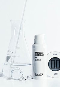 Crema Hydrafast 24h in un contenitore cilindrico bianco, un becher di vetro con liquido, sfere trasparenti e un timer digitale con pulsante di ripristino.