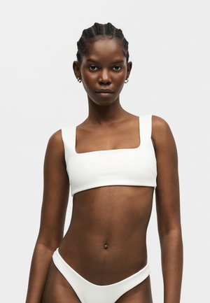 Donna con capelli intrecciati che indossa un top bikini bianco con scollo quadrato e slip abbinati, in piedi su uno sfondo chiaro e uniforme.