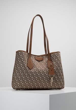 Bruine DKNY draagtassen met herhalend logopatroon, dubbele schouderriemen, leren label en een klein gouden sleutelbedel, tentoongesteld op een wit voetstuk.