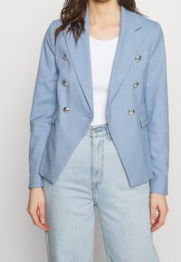 Ljusblå dubbelknäppt blazer med metallknappar, skräddarsydd passform, slagkrage och två framfickor. Bärs över en vit topp.