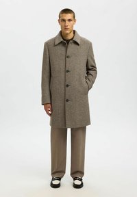 Soprabito marrone in tweed con collare, chiusura con bottoni e tasche laterali, indossato sopra un maglione marrone e pantaloni beige. Scarpe in bianco e nero.