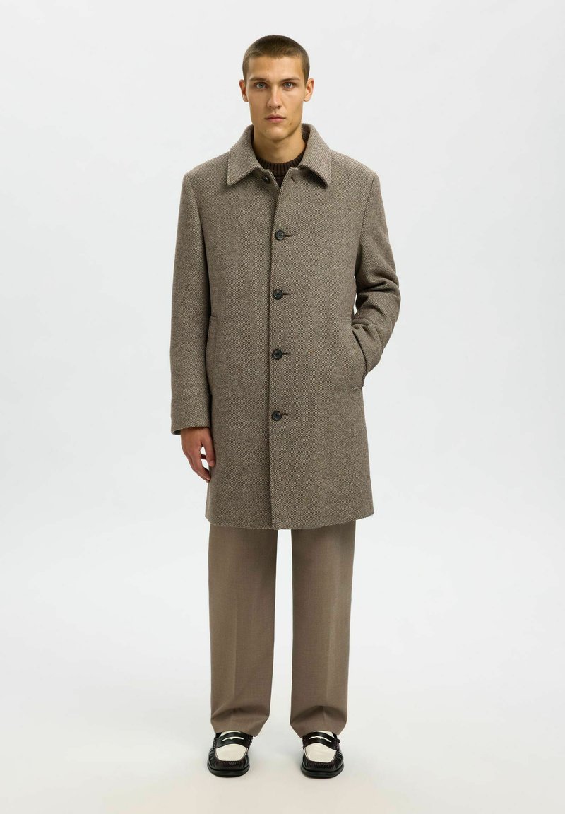 Soprabito marrone in tweed con collare, chiusura con bottoni e tasche laterali, indossato sopra un maglione marrone e pantaloni beige. Scarpe in bianco e nero.