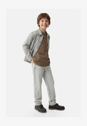 Ragazzo sorridente che indossa una giacca di jeans grigio chiaro e jeans, una t-shirt marrone e scarpe nere, in piedi con una mano in tasca su sfondo bianco.