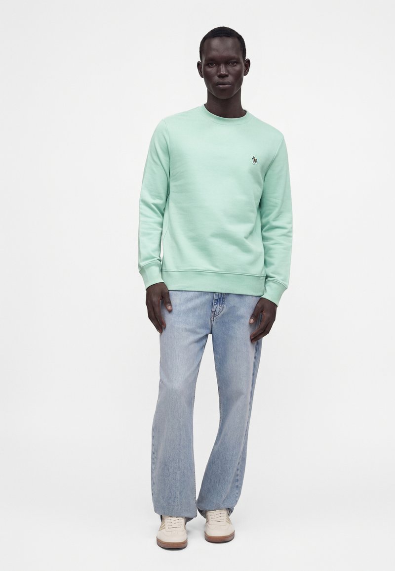 Homme debout portant un sweat-shirt vert menthe, un jean bleu clair et des baskets blanches sur un fond blanc uni.