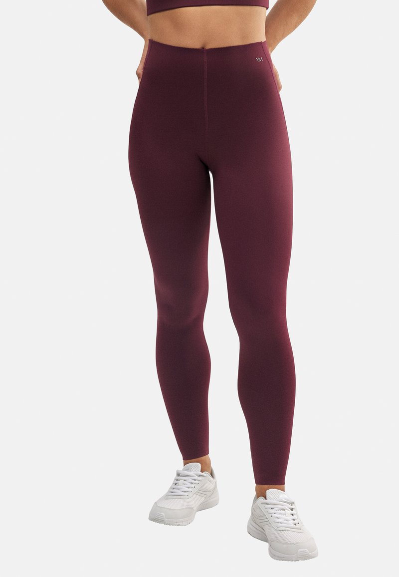 Leggings color burdeos con textura suave, cintura alta y diseño ajustado, combinados con zapatillas deportivas blancas. Detalle de costura simple visible.