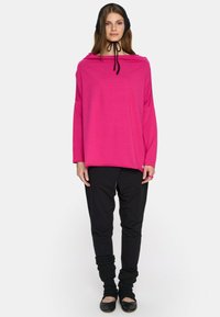 Na różowawym oversizedowym topie z drapowanym dekoltem, długimi rękawami i czarnym sznurkiem. Połączony z czarnymi zwężającymi się spodniami i prążkowanymi czarnymi ocieplaczami na nogi.