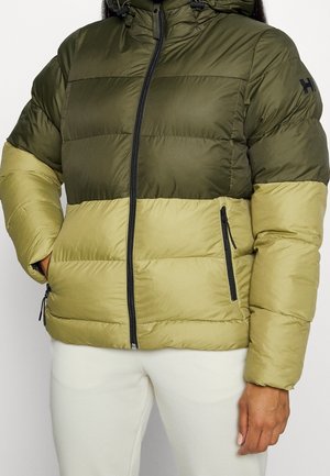 Mujer con chaqueta acolchada bicolor en verde oliva y verde claro con cremallera frontal y bolsillos laterales, combinada con pantalones blancos.