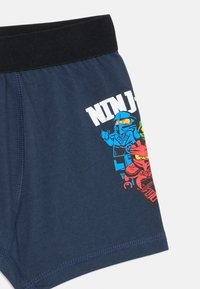 Blaue Baumwollshorts mit einem schwarzen Bund, mit einem bunten Grafikdesign von Ninja-Figuren an der Seite. Glatte Textur.