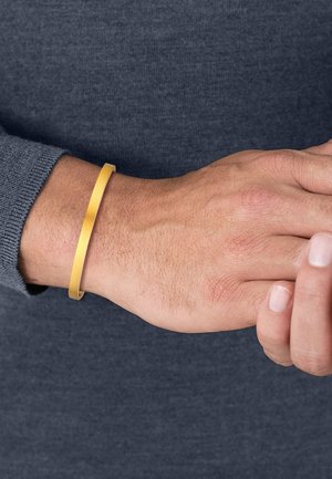 Bracelet en métal doré sur un poignet, présentant un design lisse et plat avec une texture brossée et des bords arrondis.