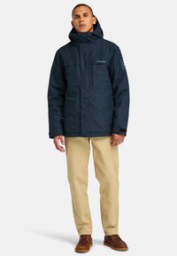 Marineblauwe waterdichte jas met een capuchon, voorzien van een rits aan de voorkant, twee borstzakken en logo-opdruk aan de linkerkant. Beige broek en bruine schoenen.