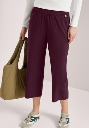 Femme portant un pantalon court bordeaux, un haut à manches longues blanc, des baskets blanches et vertes, tenant un sac cabas matelassé vert olive.
