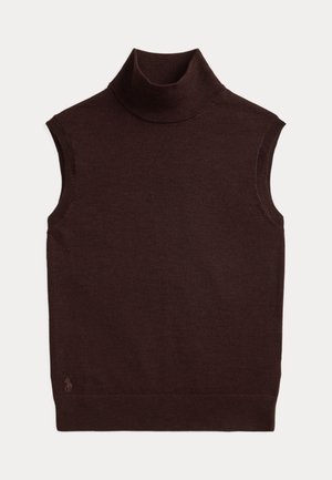 Frau steht mit der Hand in der Tasche, trägt einen grauen Anzug, einen dunklen Rollkragenpullover, einen breiten Gürtel mit großen silbernen Schnallen und braune spitze Schuhe.