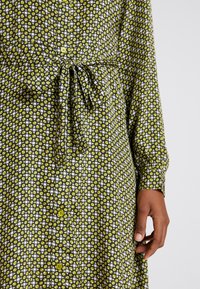 Long-sleeve shirt dress made of patterned fabric featuring green and white geometric shapes, cinched at the waist with a tie, and button details.