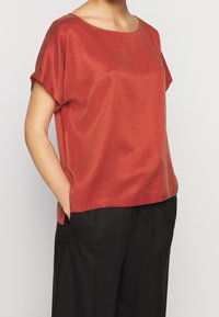 Rustfarvet kortærmet top med en bred halsudskæring og glat tekstur. Kombineret med sorte, løstsiddende bukser. Simple og minimalistisk design.