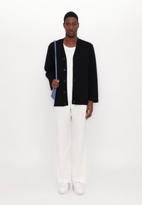 Veste noire à boutonnière avec une coupe décontractée, portée sur un haut blanc, associée à un pantalon crème et des baskets blanches, tenant un sac à bandoulière bleu.